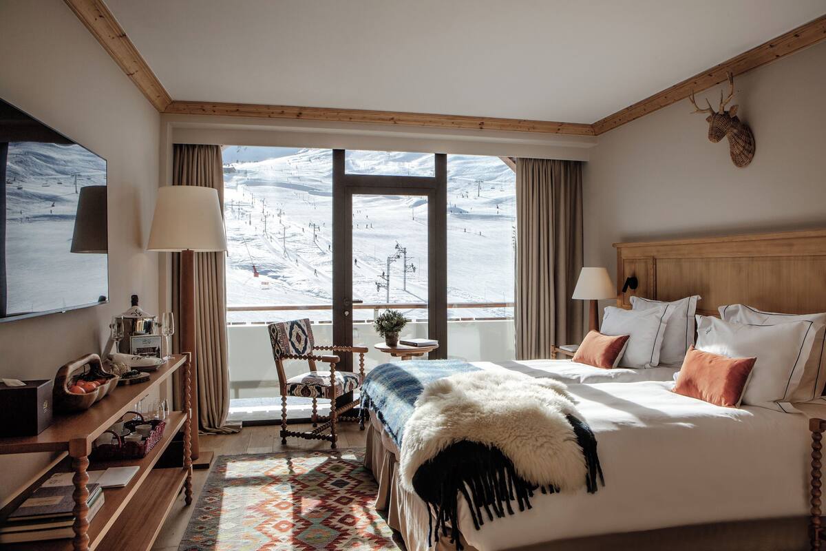 Ein Hotelzimmer mit einem großen Bett, einem hölzernen Kopfteil, einem Schreibtisch mit Lampe, einem Sessel, einem Fernseher und Blick auf eine verschneite Landschaft.