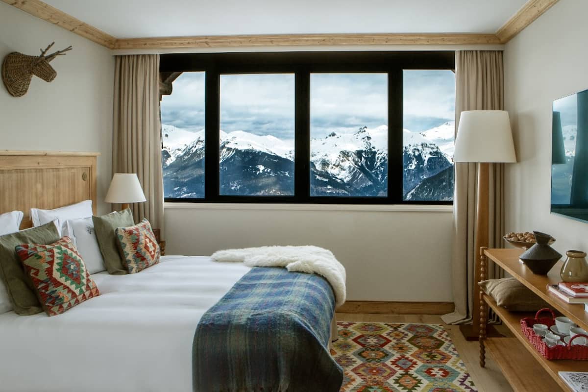 Ein Hotelzimmer mit Bett, Blick auf schneebedeckte Berge, einem Holzkopfstück, einer Lampe und einem kleinen Tisch mit einer Vase.