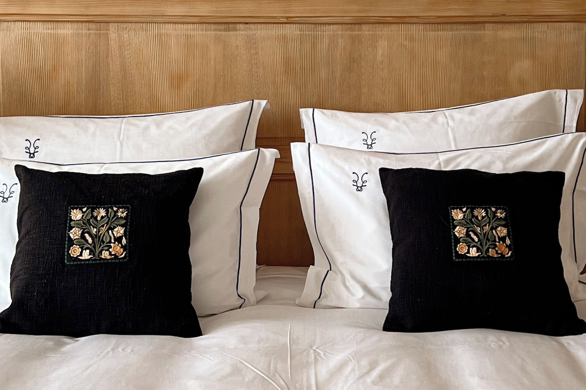 A bed with white bedding and two black pillows with floral embroidery, a wooden headboard, and a fluffy white throw.
