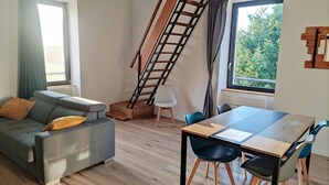 A modern living room with a grey sofa, wooden floor, a dining table with chairs, a wooden ladder, and a window with a view of greenery. - Appartement Duplex Dans une Ancienne Usine (Beaupréau-en-Mauges)