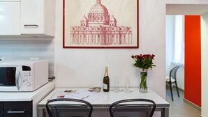 Una cocina con armarios blancos, un microondas, una mesa de comedor con sillas, un cuadro de un edificio, una botella de champán y un florero con flores.