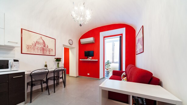Una sala de estar moderna con una pared de acento roja, un sofá blanco, una zona de comedor con mesa y sillas, y una cocina con microondas y un reloj.