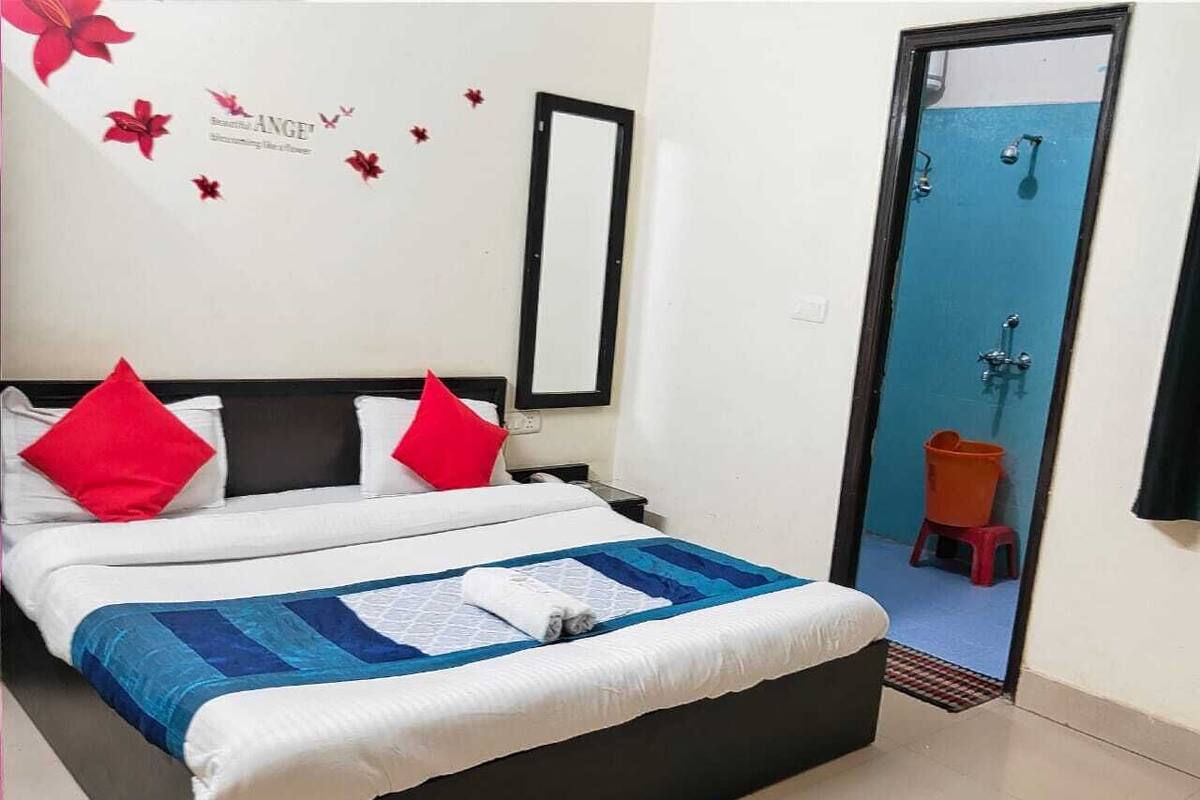Un lit bien fait, recouvert d’une couette bleue et blanche, avec des oreillers rouges et une couette blanche. On y voit une tête de lit noire, un miroir fixé au mur et une porte donnant sur une salle de bain avec une douche bleue.