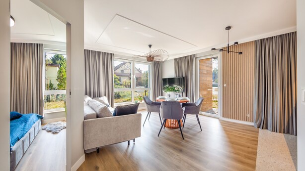 A modern living room with a sofa, dining table, and chairs, large windows with curtains, and a TV.