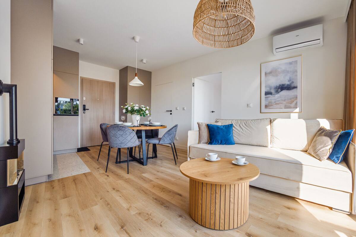Una sala moderna con suelo de madera, una mesa redonda de centro, un sofá blanco con cojines azules y beige, y un comedor con mesa y sillas.