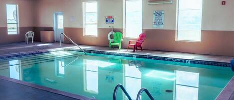 An indoor swimming pool with clear water, a diving board, and three chairs near the windows.