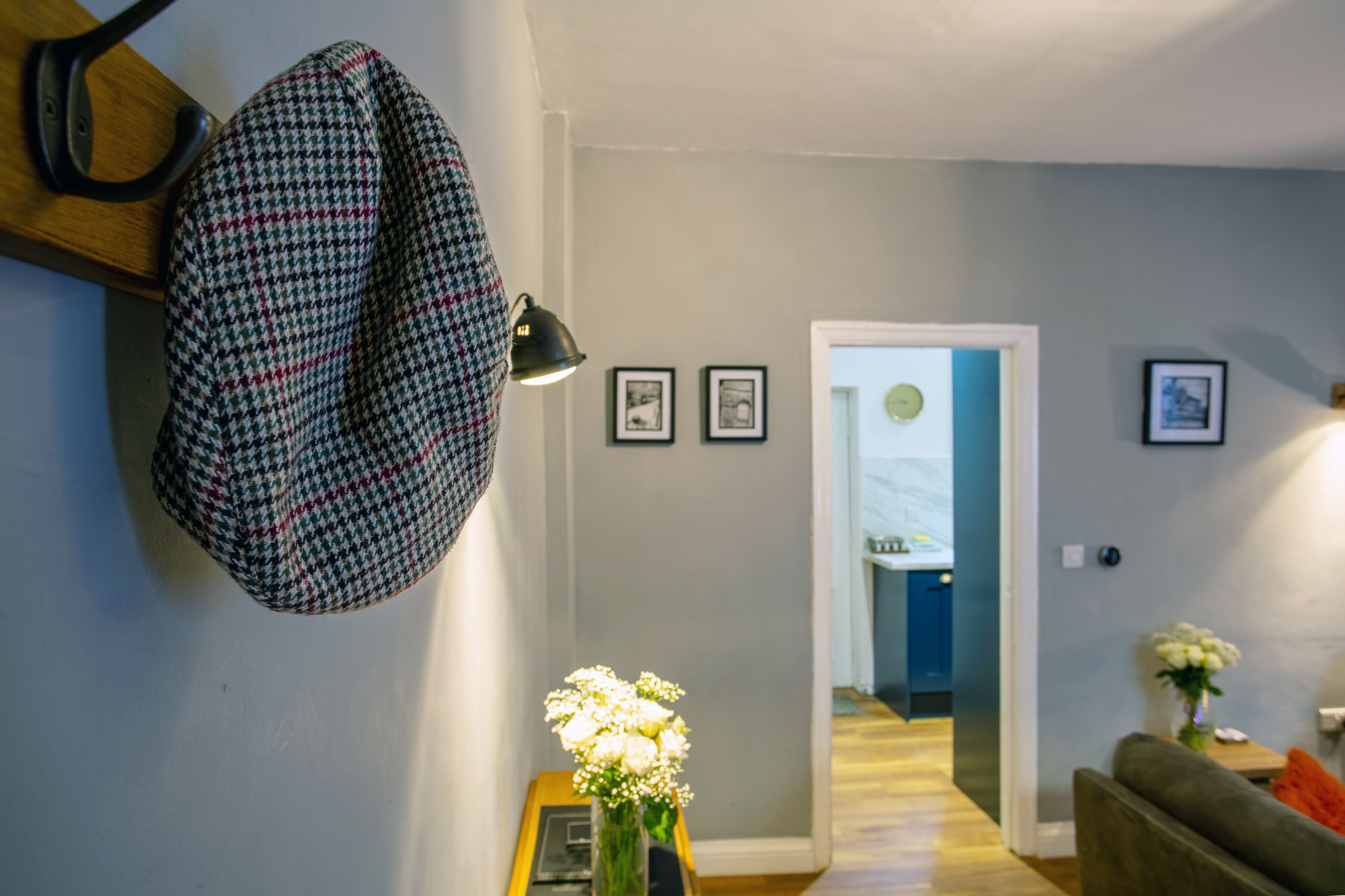 A hallway with a houndstooth-patterned coat hanging on a hook, a vase with white flowers, and framed pictures on the wall.