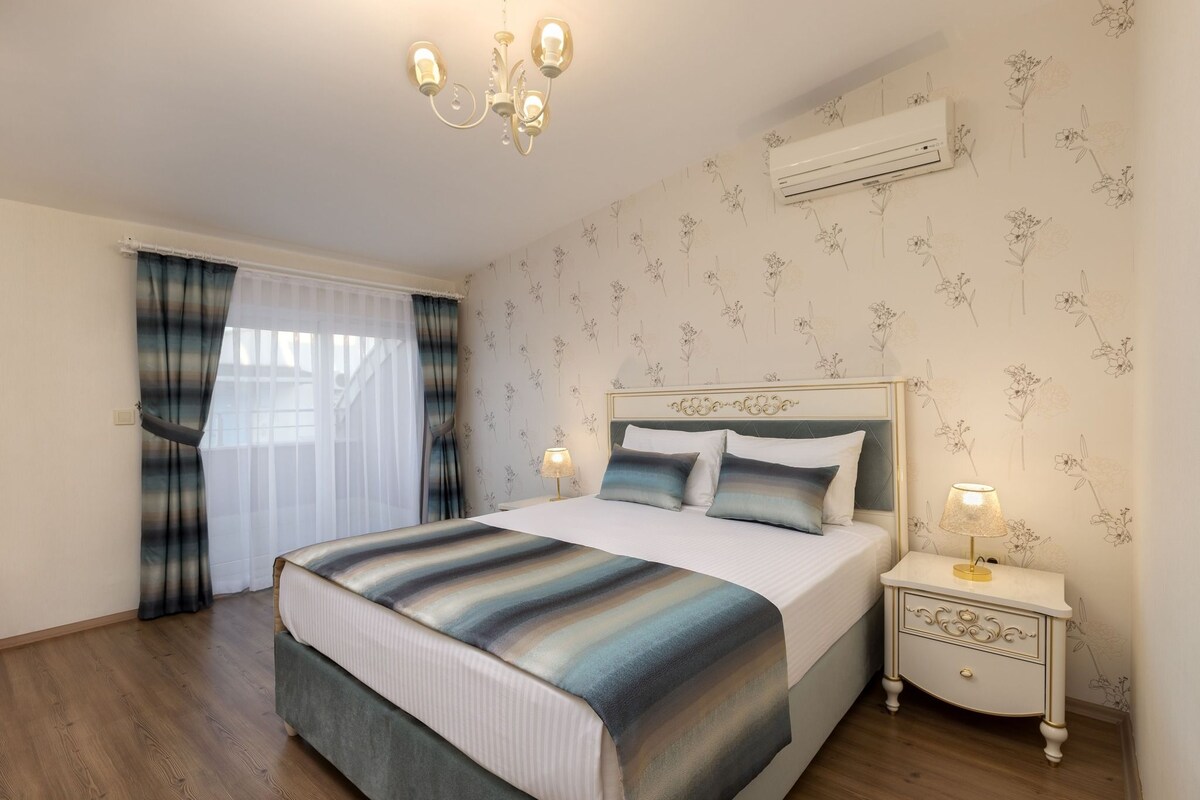 A bedroom with a bed, bedside tables, a chandelier, and an air conditioning unit.