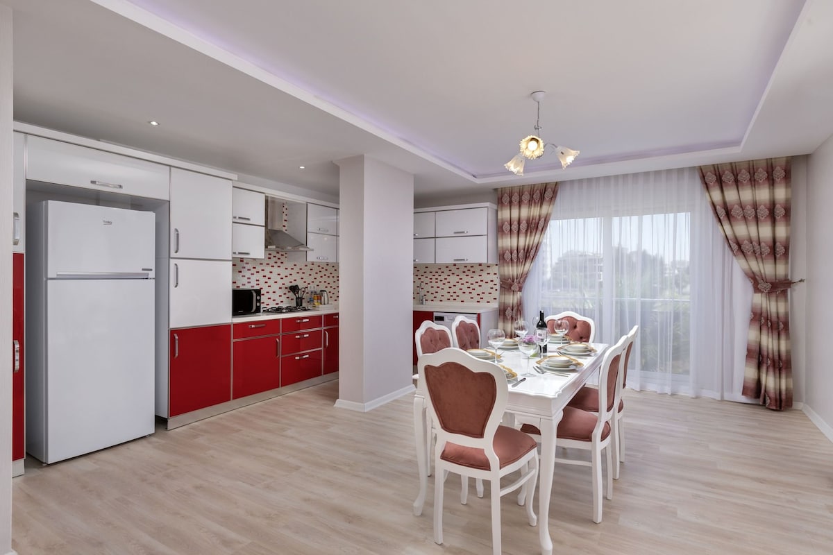 Une cuisine moderne avec des meubles blancs et rouges, un coin repas avec une table et des chaises blanches, et une grande fenêtre avec des rideaux.