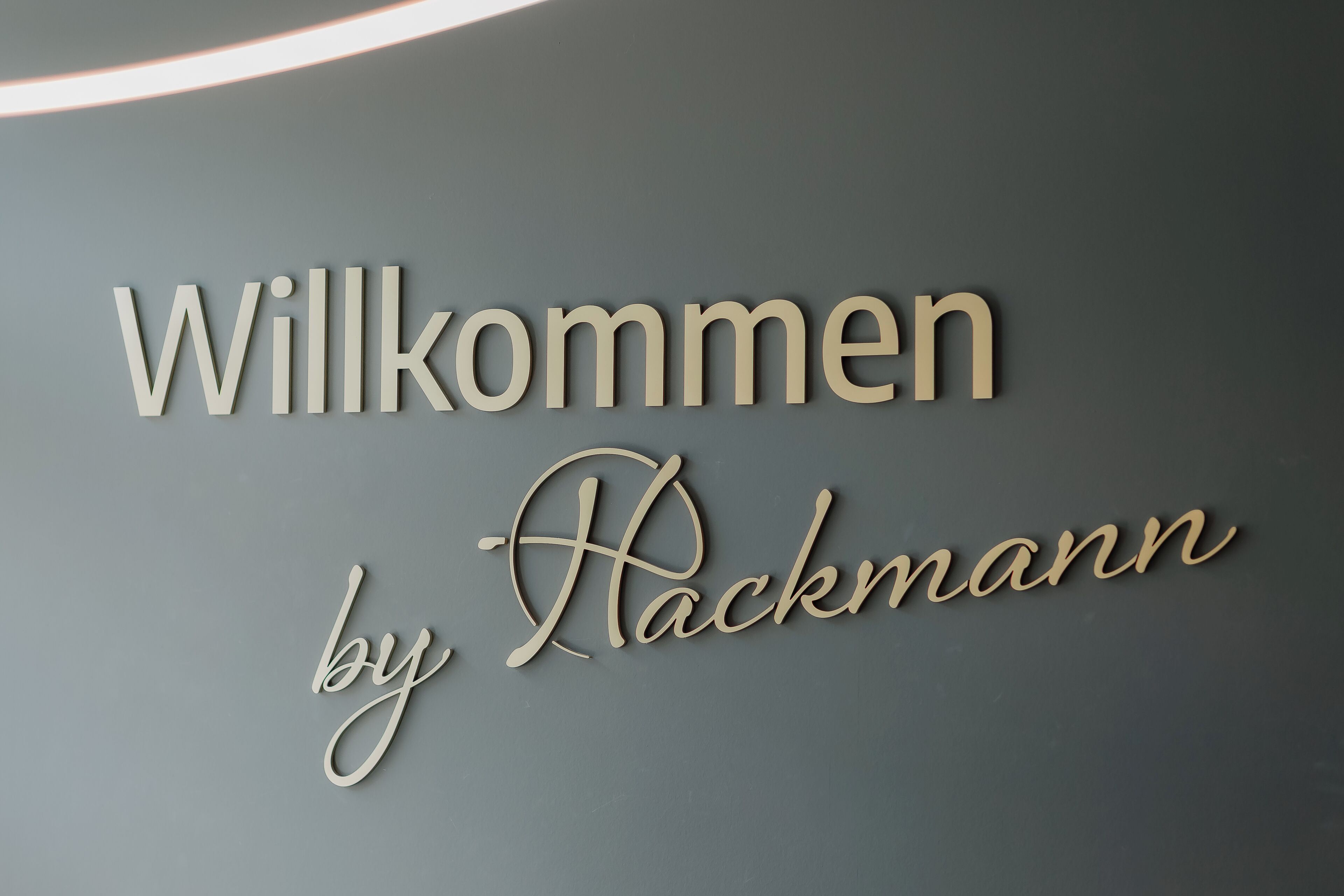 Letrero de bienvenida con el texto 'Willkommen by Rackmann' sobre una pared gris.