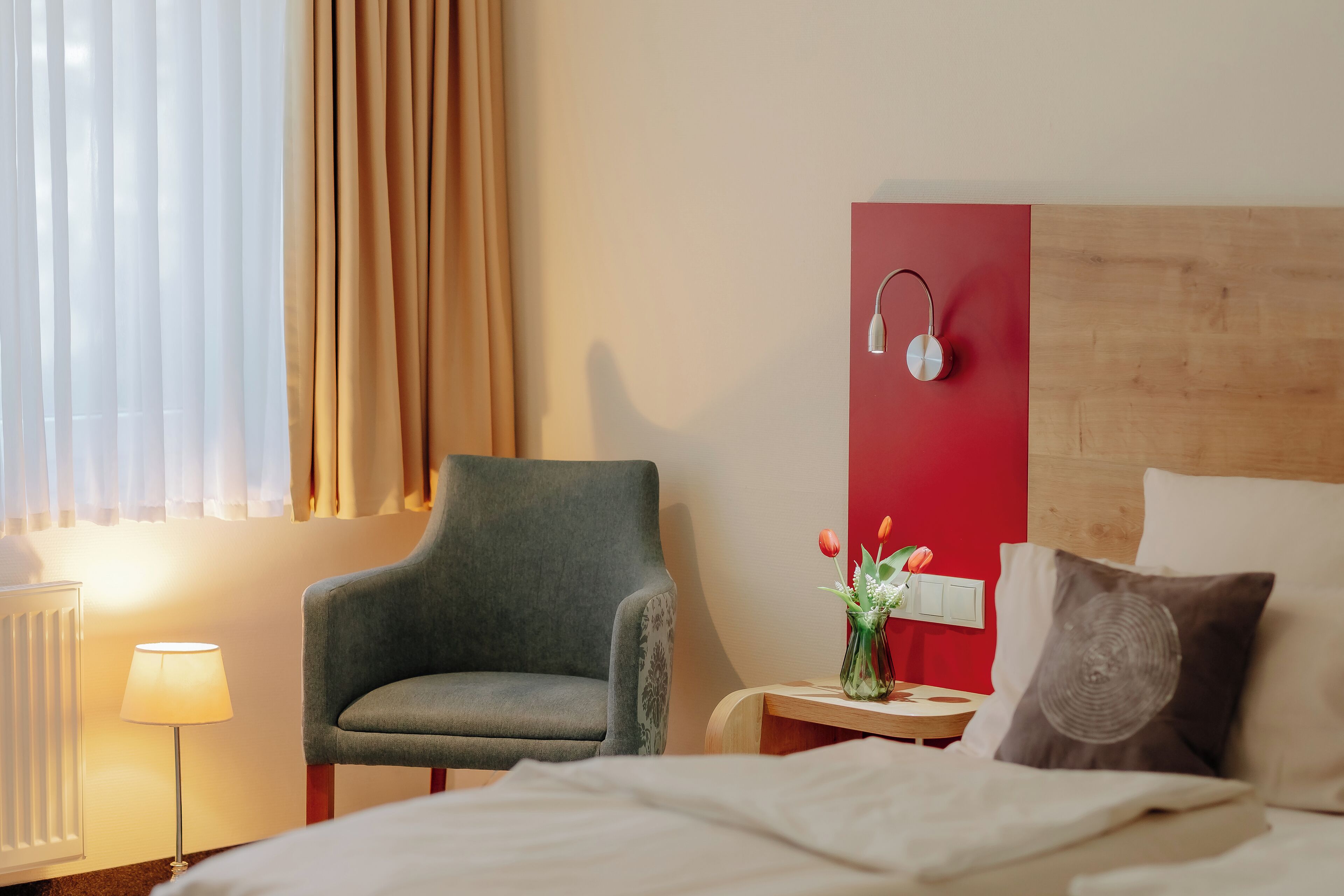 Habitación de hotel con una cama, una silla, una mesita de noche con lámpara y un altavoz en la pared.