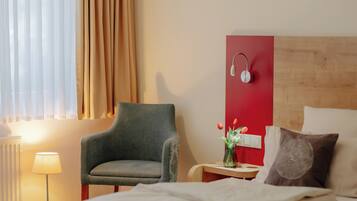 A hotel room with a bed, a chair, a bedside table with a lamp, and a wall-mounted speaker.