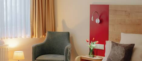Habitación de hotel con una cama, una silla, una mesita de noche con lámpara y un altavoz en la pared.