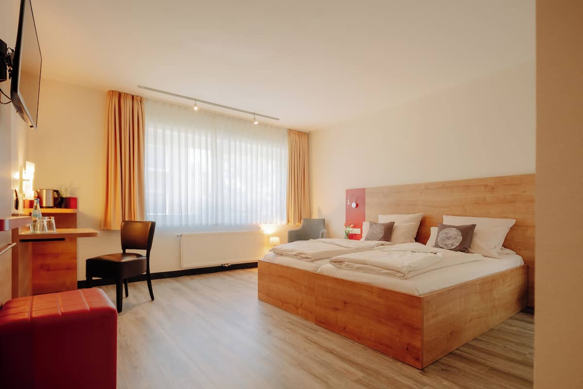 Quarto de hotel moderno com cama de madeira, escrivaninha com cadeira e uma janela grande com cortinas.