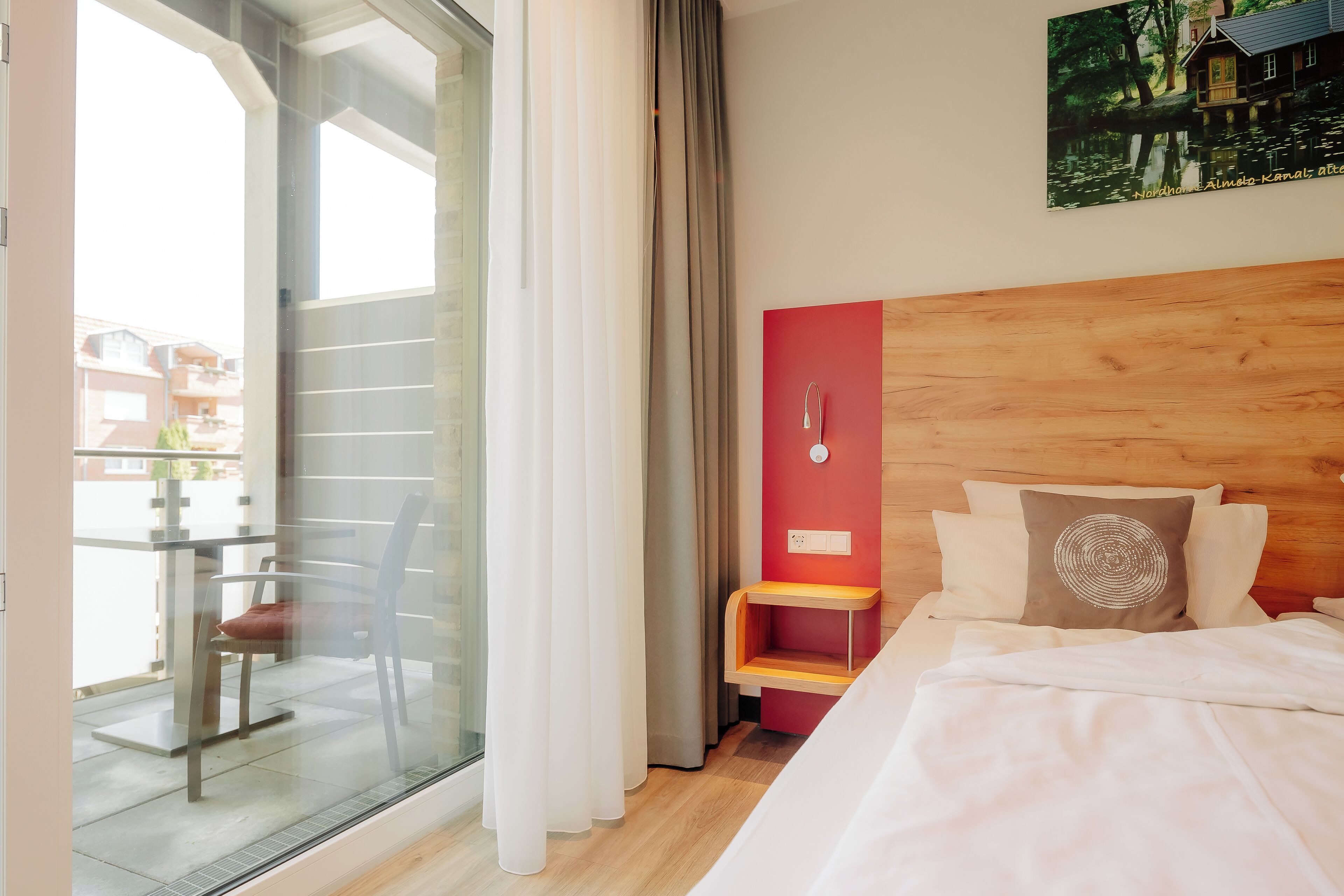Ein Hotelzimmer mit einem Bett, einem kleinen Holz-Nachttisch, einer roten Akzentwand und Blick auf ein Gebäude durch eine Glastür.