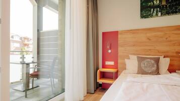 Ein Hotelzimmer mit einem Bett, einem kleinen Holz-Nachttisch, einer roten Akzentwand und Blick auf ein Gebäude durch eine Glastür.