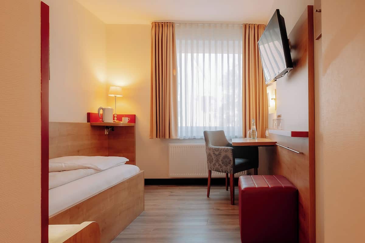 Quarto de hotel pequeno com cama, escrivaninha, cadeira, TV fixada na parede e janela com cortinas.