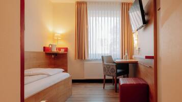 Habitación de hotel compacta con cama, escritorio, silla, televisor montado en la pared y ventana con cortinas.