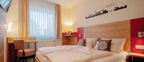 A hotel room with a bed, desk, chair, and a cityscape wall decoration.