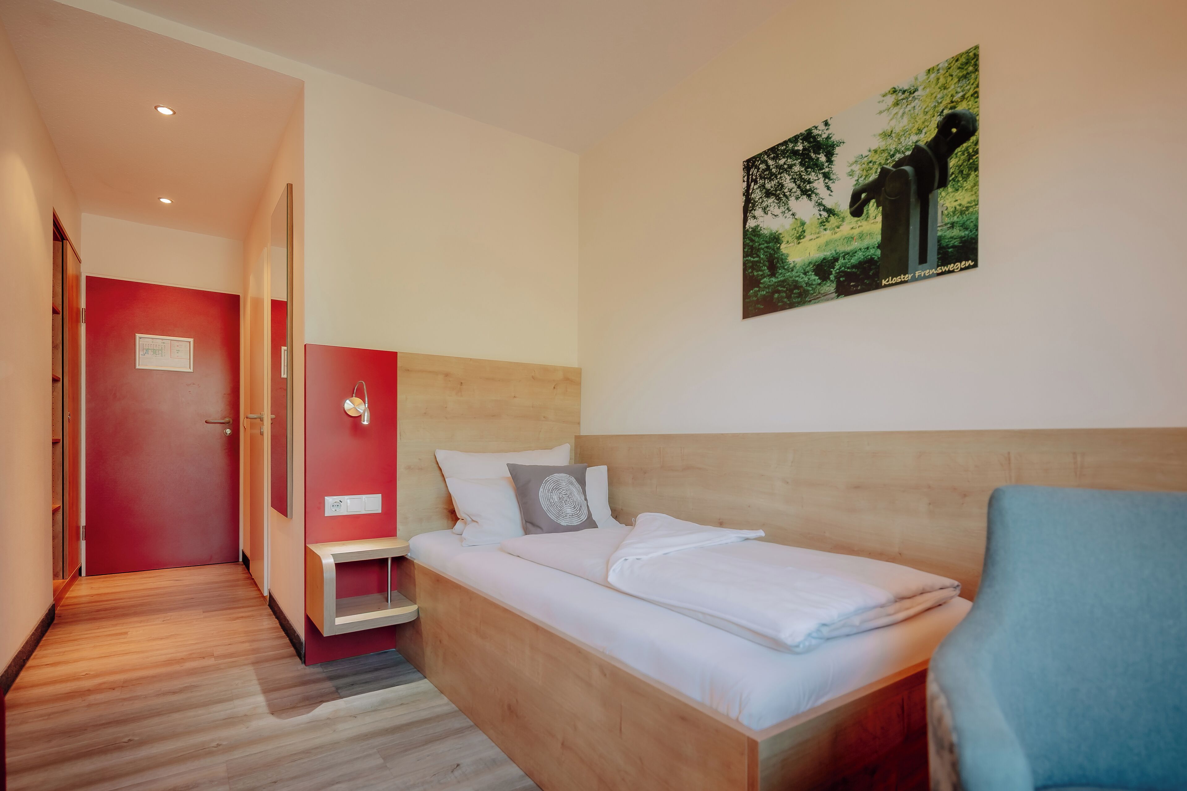 A hotel room with a wooden bed, a red wall, a small bedside table, and a picture of a tree.