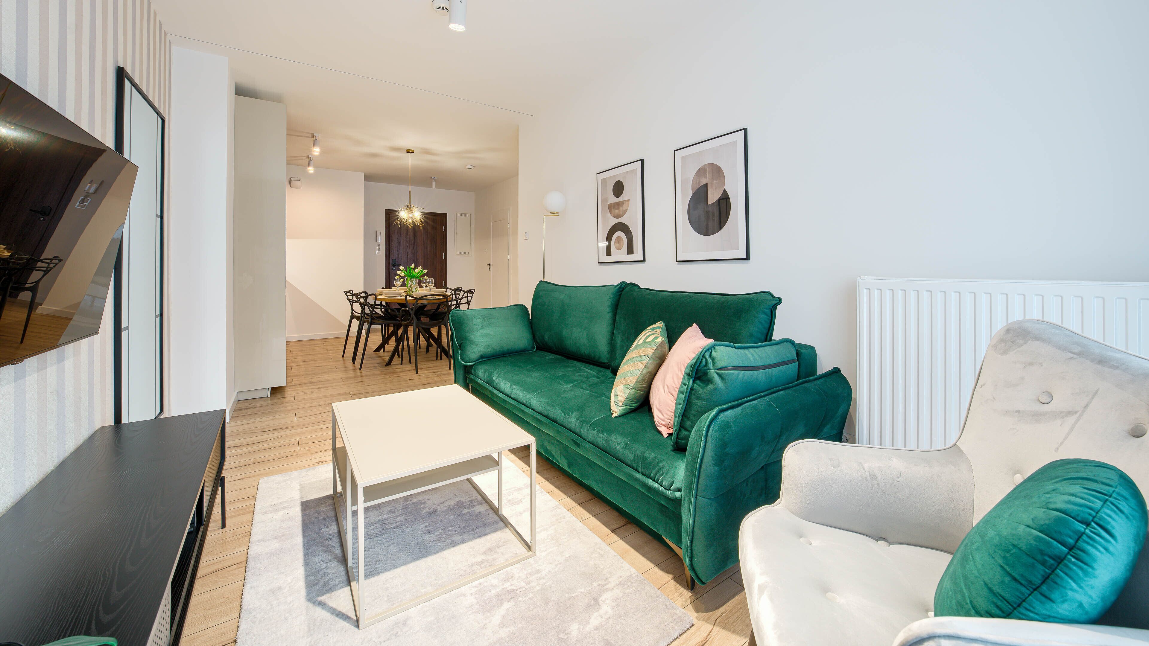 Ein modernes Wohnzimmer mit einem grünen Sofa, einem weißen Sessel und einem Essbereich im Hintergrund.