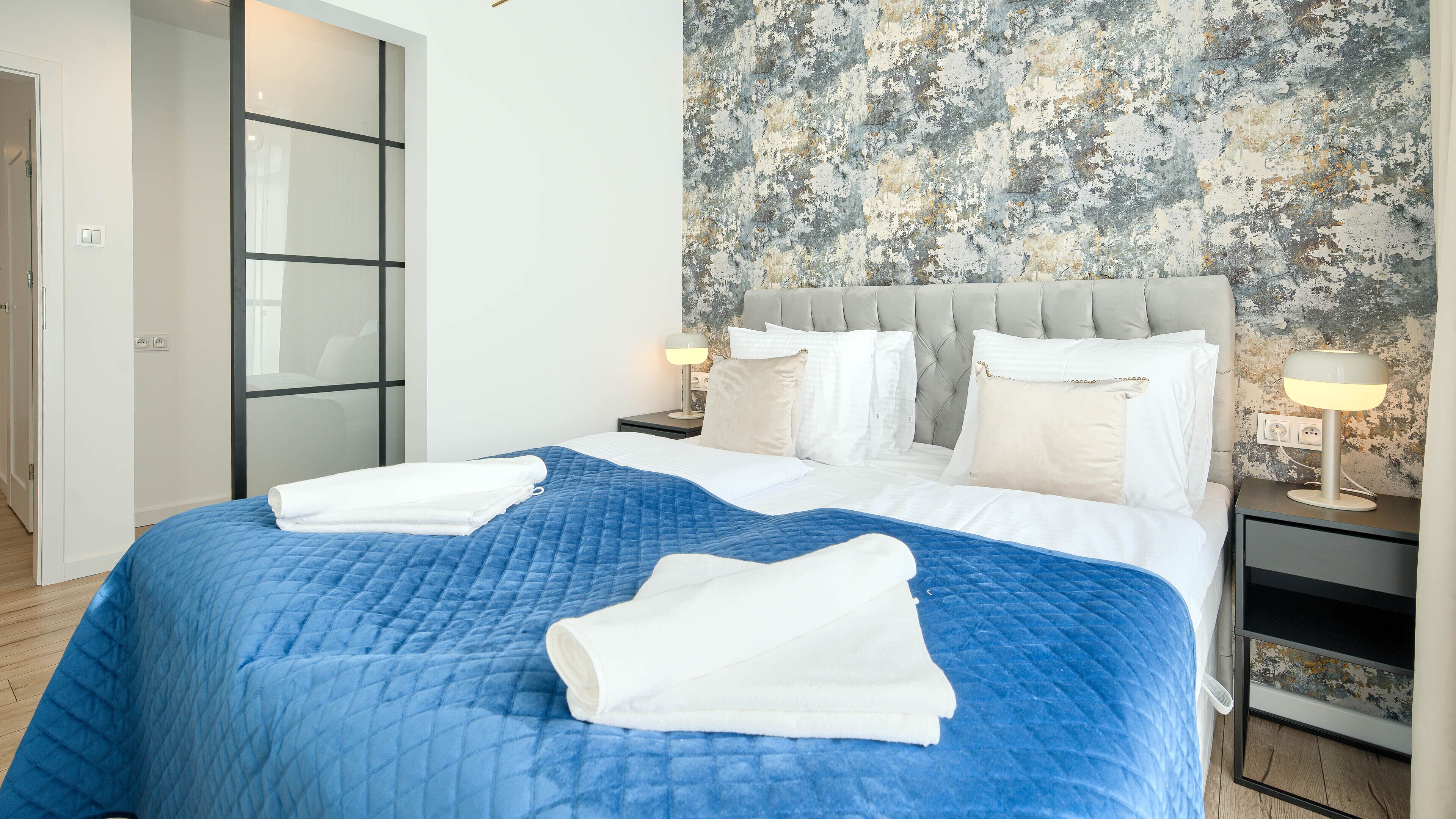 A neatly made bed with a blue quilt, white pillows, and folded white towels. There is a bedside table with a lamp and a power outlet. The room has a modern design with a textured wall and a sliding door.