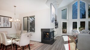 En moderne stue med et glasbord, hvide stole, pejs og store vinduer.