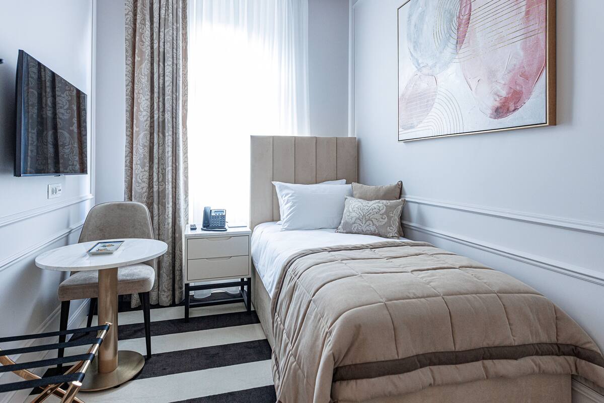 Une chambre d’hôtel bien rangée, avec un lit, une table de chevet, une chaise et une grande fenêtre avec des rideaux.