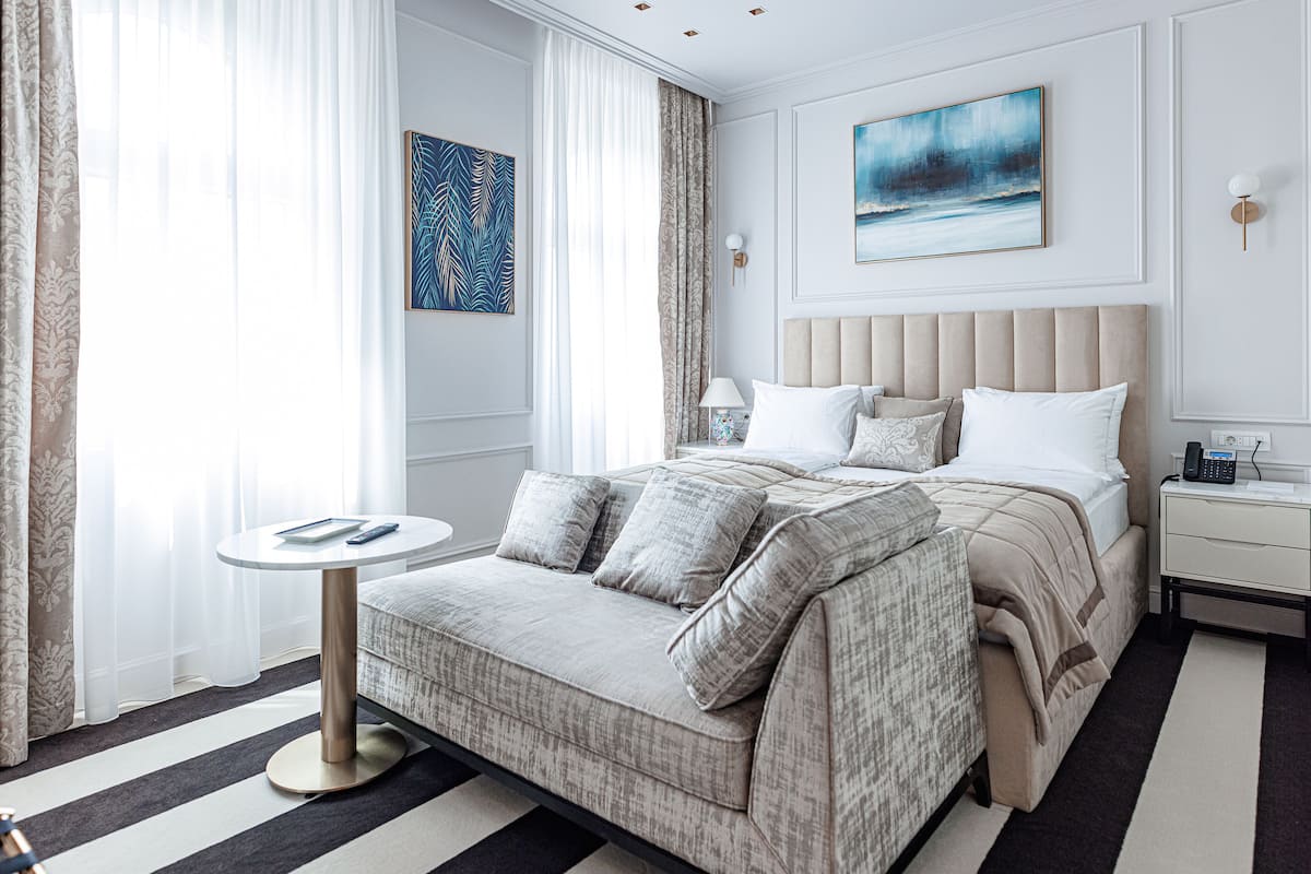 Une chambre d’hôtel moderne avec un grand lit, une table ronde et une vue sur la ville à travers de fines rideaux.