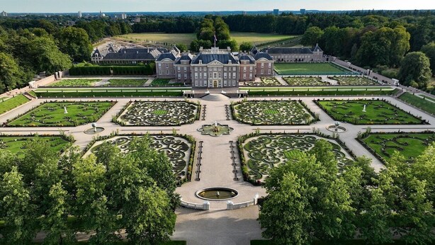 Ein großer, symmetrischer Garten mit einem zentralen Brunnen, umgeben von gepflegten Rasenflächen und einem imposanten Gebäude mit einem markanten Eingang.