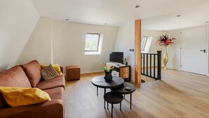 Ein modernes Wohnzimmer mit einem braunen Sofa, einem kleinen runden Tisch und einem Fernseher auf einem Holzständer. Ein Fenster lässt natürliches Licht herein und bietet Blick auf eine grüne Landschaft.