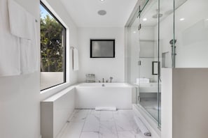 Ein modernes Badezimmer mit Badewanne, Duschkabine aus Glas und einem großen Fenster mit Blick auf Bäume.