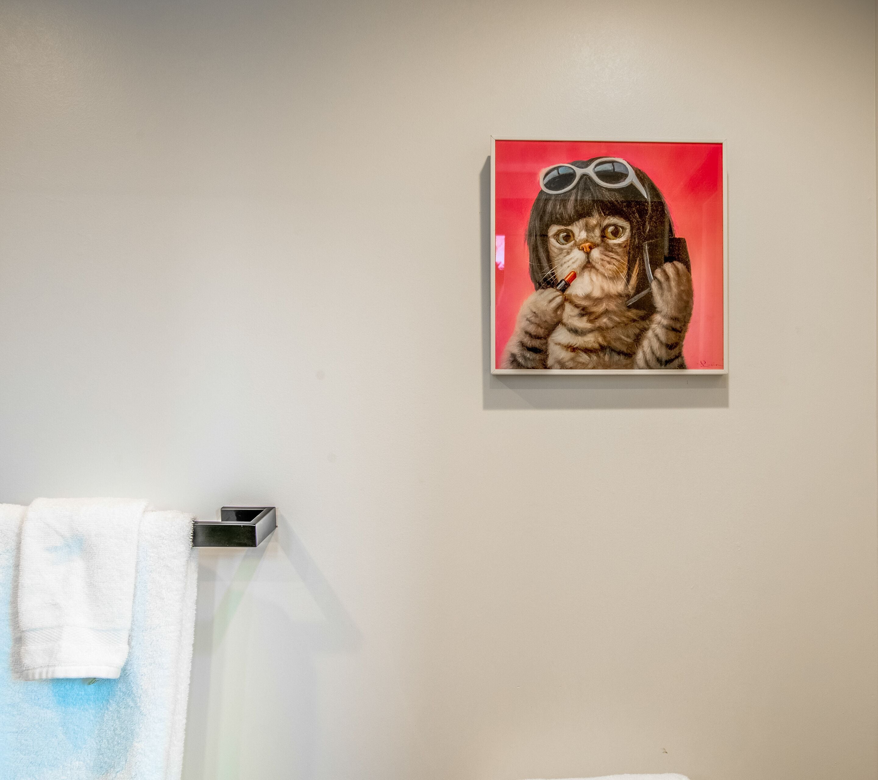 Un baño con un perchero que sostiene una toalla blanca, una obra de arte enmarcada de un gato con gafas de sol, y un fondo rojo.