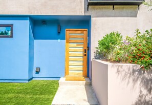 A modern building with a blue exterior, a wooden door, a small window, and a concrete pathway leading to the entrance. - Stylish Sherman Oaks Townhouse W/ 3 Master Suites (Van Nuys)