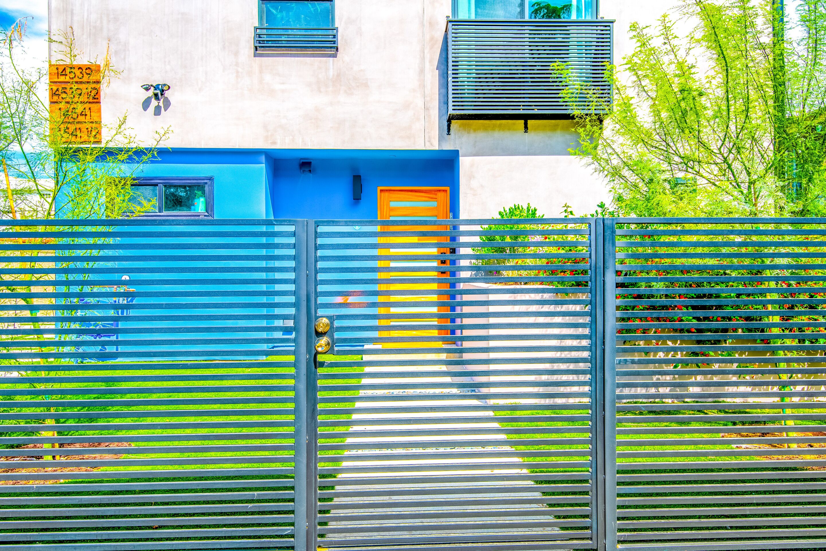 Une entrée fermée par un portail, avec une porte bleue et jaune, un portail en métal et un bâtiment dont la façade et les fenêtres sont de couleur claire.