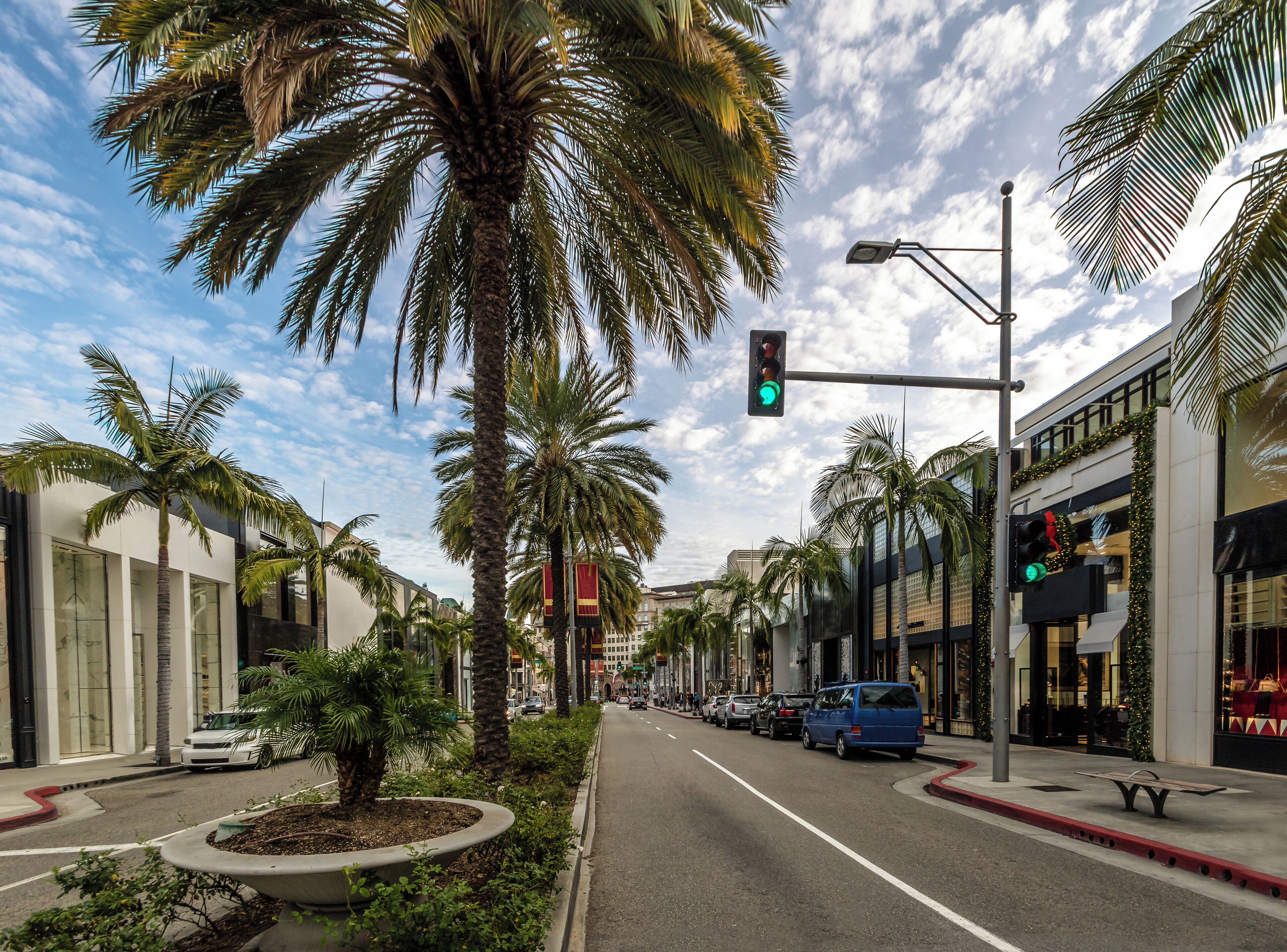 A street lined with palm trees and modern buildings, a green traffic light, and parked cars.