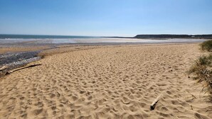 Een breed zandstrand met zachte golven, een heldere lucht en heuvels in de verte.