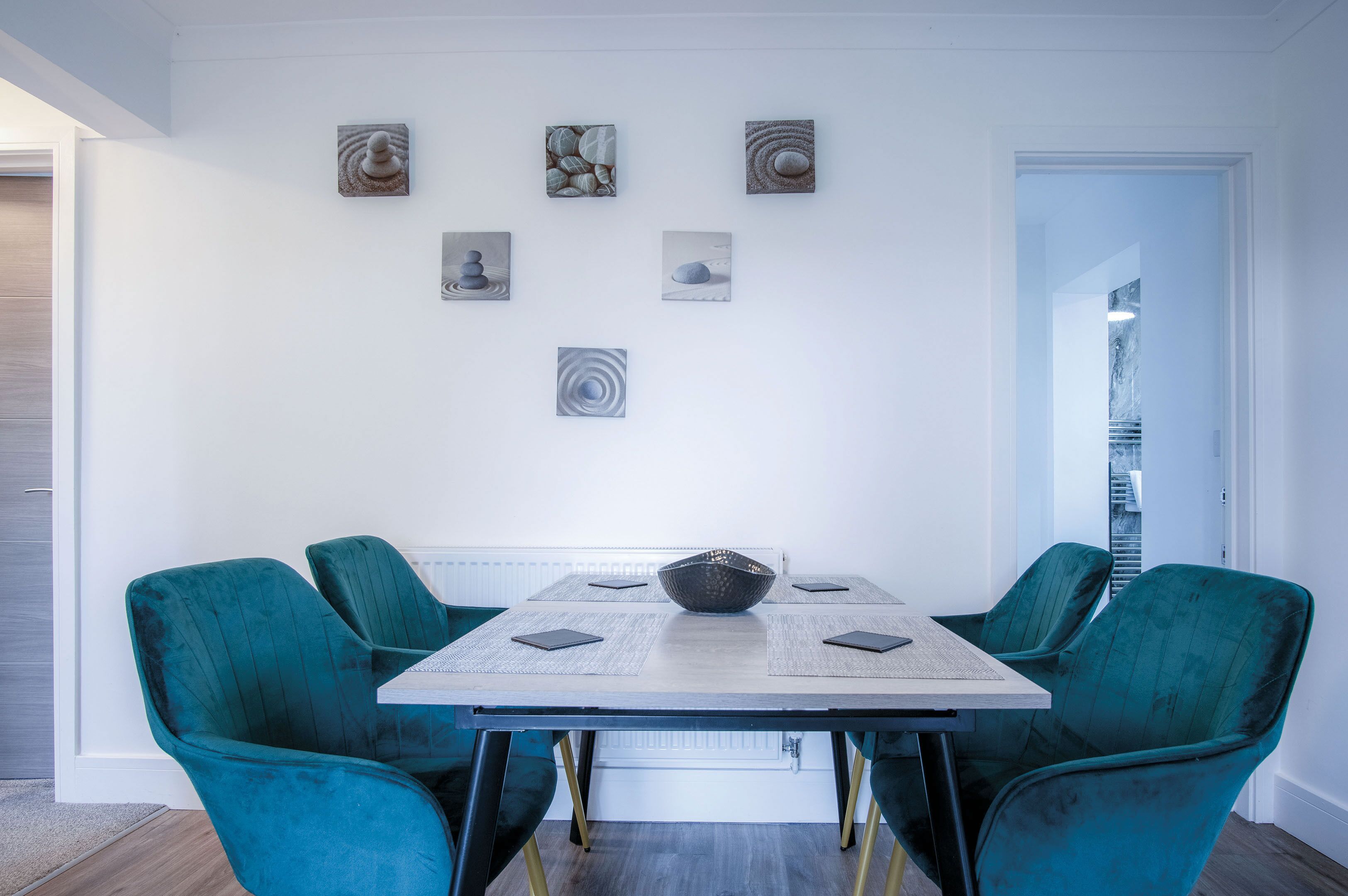 A dining area with a table and four teal chairs, a bowl in the center, and six framed artworks on the wall.