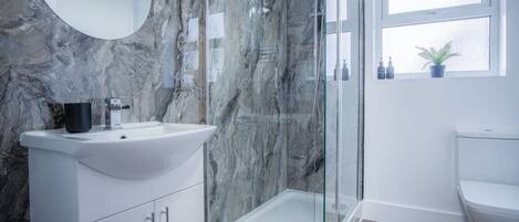 Une salle de bain moderne avec un mur en marbre, une cabine de douche en verre, un lavabo avec un miroir et une fenêtre agrémentée d’une plante.