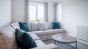A modern living room with a sectional sofa, a round coffee table, and a view of the outdoors through large windows. - Gower Hideaway - 3 Bed - Ocean Breeze (Swansea)