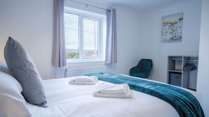 A neatly made bed with white linens and two folded towels, a window with curtains, a teal chair, and a painting on the wall. - Gower Hideaway - 3 Bed - Ocean Breeze (Swansea)