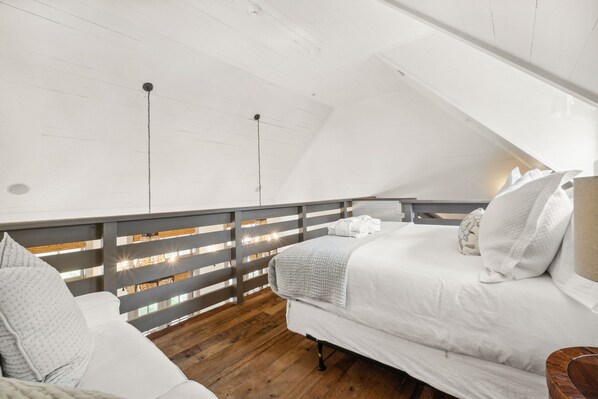 Un dormitorio con techo inclinado, piso de madera y cama con ropa blanca.