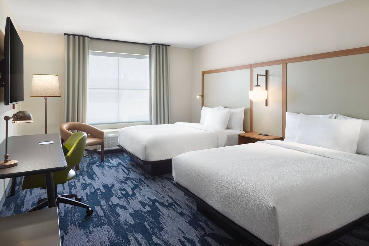 A hotel room with two beds, a desk, a chair, a television, and a large window with curtains.