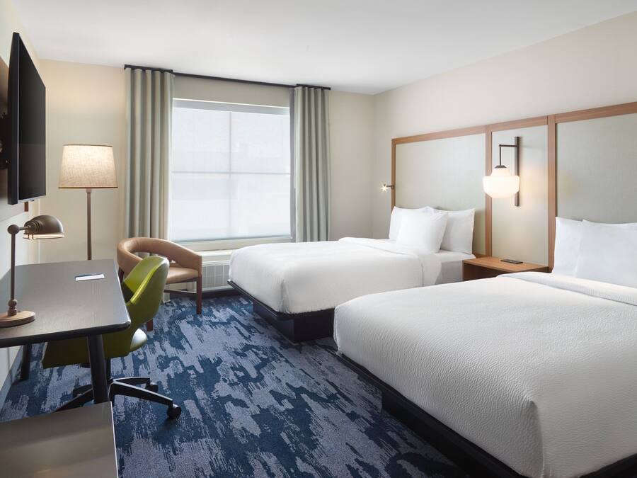 A hotel room with two beds, a desk, a chair, a television, and a large window with curtains.