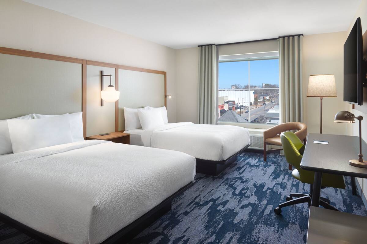 A hotel room with two beds, a desk, a chair, a TV, and a large window with a view of the city.