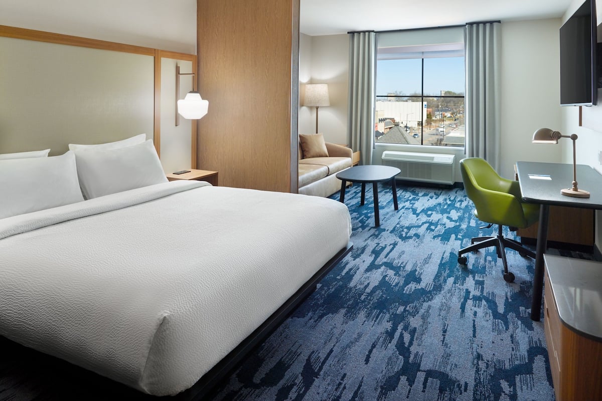 A hotel room with a large bed, a desk with a chair, a small table, a lamp, and a TV.