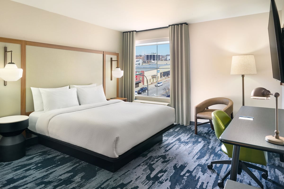 A hotel room with a large bed, a desk, a chair, a lamp, and a view of the city through the window.