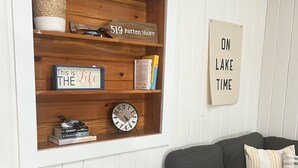 A living room with a dark grey sofa, a grey armchair, a wooden shelf with books and a clock, and a sign that reads "ON LAKE TIME". - Charming Lakehouse private yard, boat dock/wifi. (Frankling Vermont)