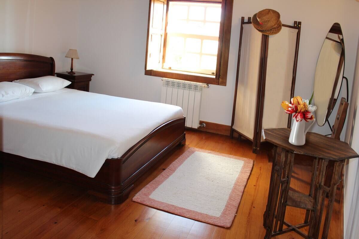 Una cama bien tendida con sábanas blancas, una mesita de noche de madera con una lámpara, un radiador, una ventana con vistas, un espejo y un jarrón con flores sobre una mesa de madera.