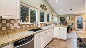 A kitchen with white cabinets, granite countertops, and a dishwasher. There are windows with views of trees, a dining area with a table and chairs, and a ceiling with recessed lighting.